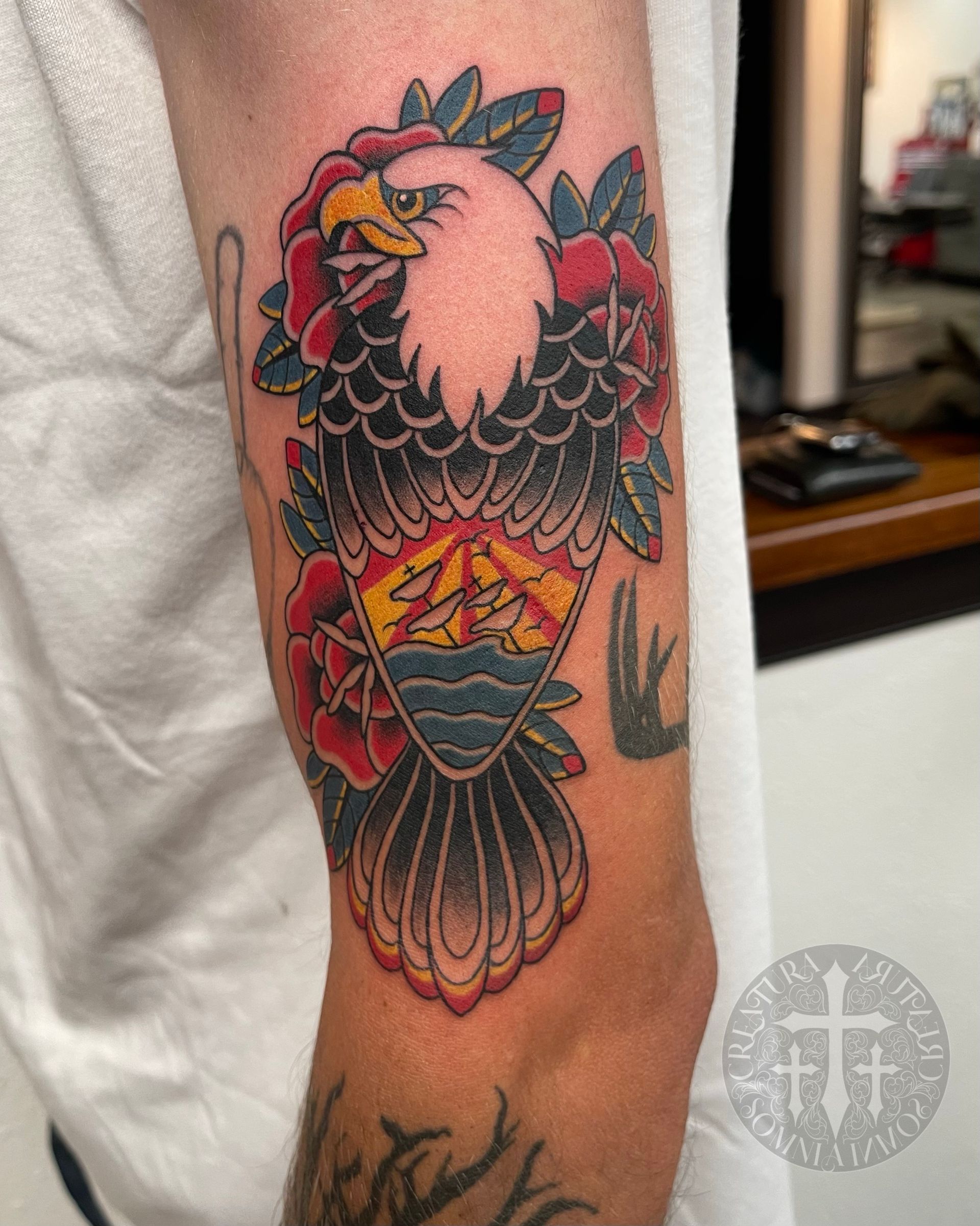 Tattoo of an eagle with red roses on arm. The eagle has wings spread, and a landscape inside.