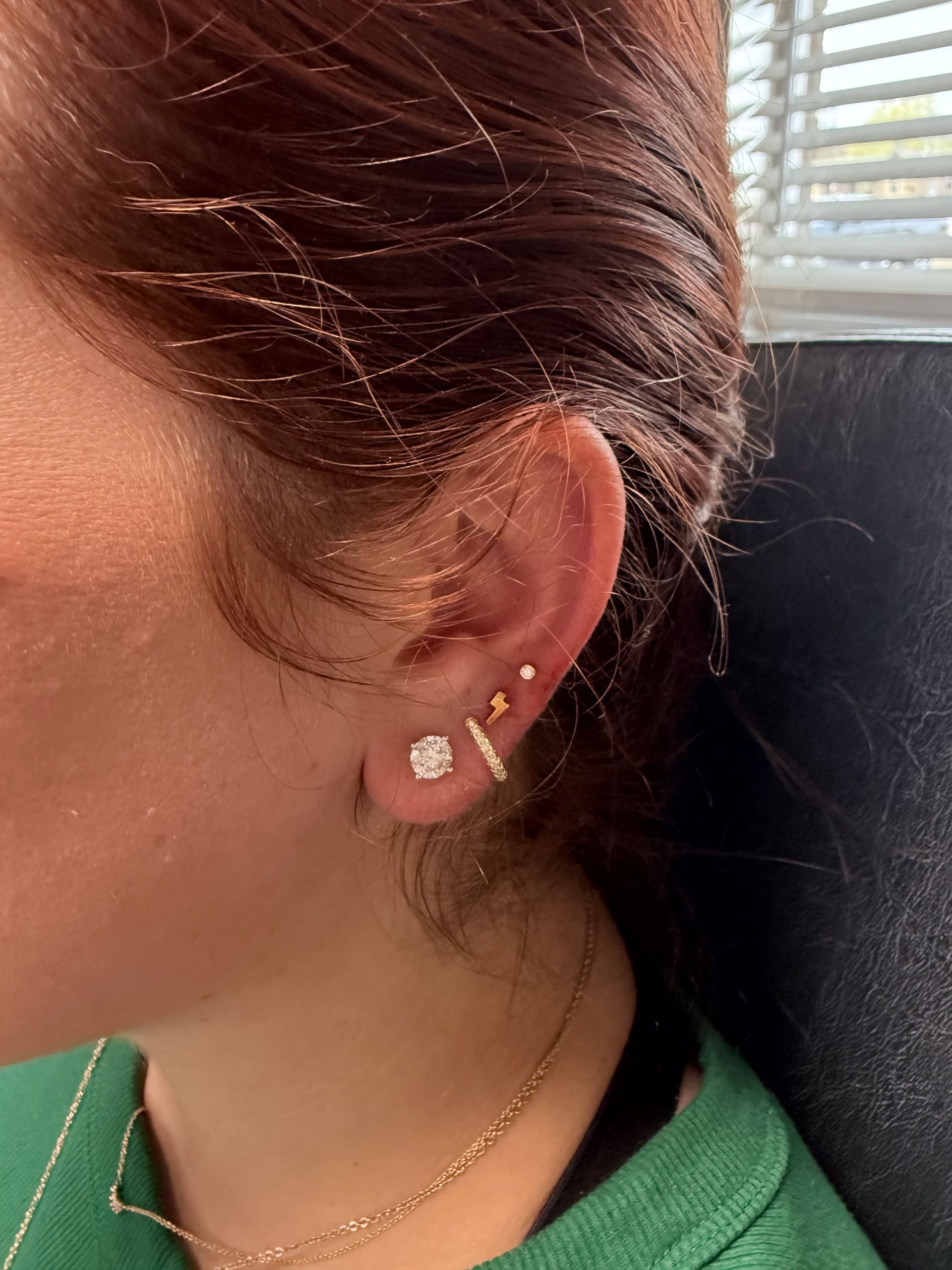 Profile view of a person's ear with multiple earrings. The person wears a green shirt and gold necklace.