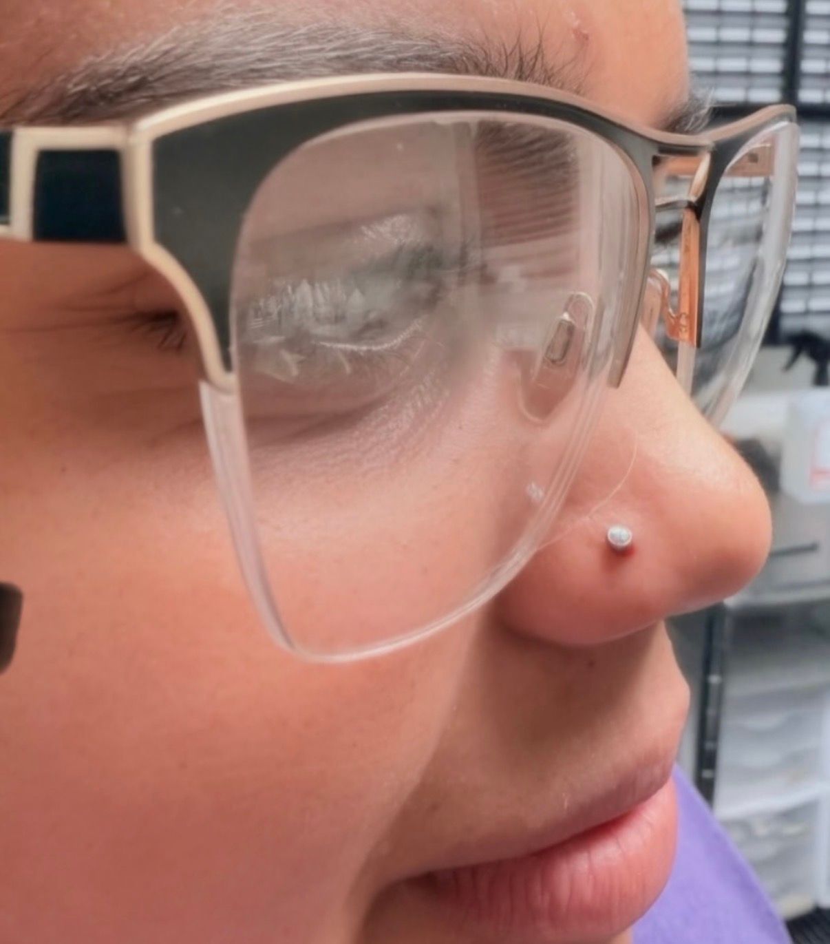 Person wearing glasses with a nose piercing. The piercing is a small stud on their right nostril.