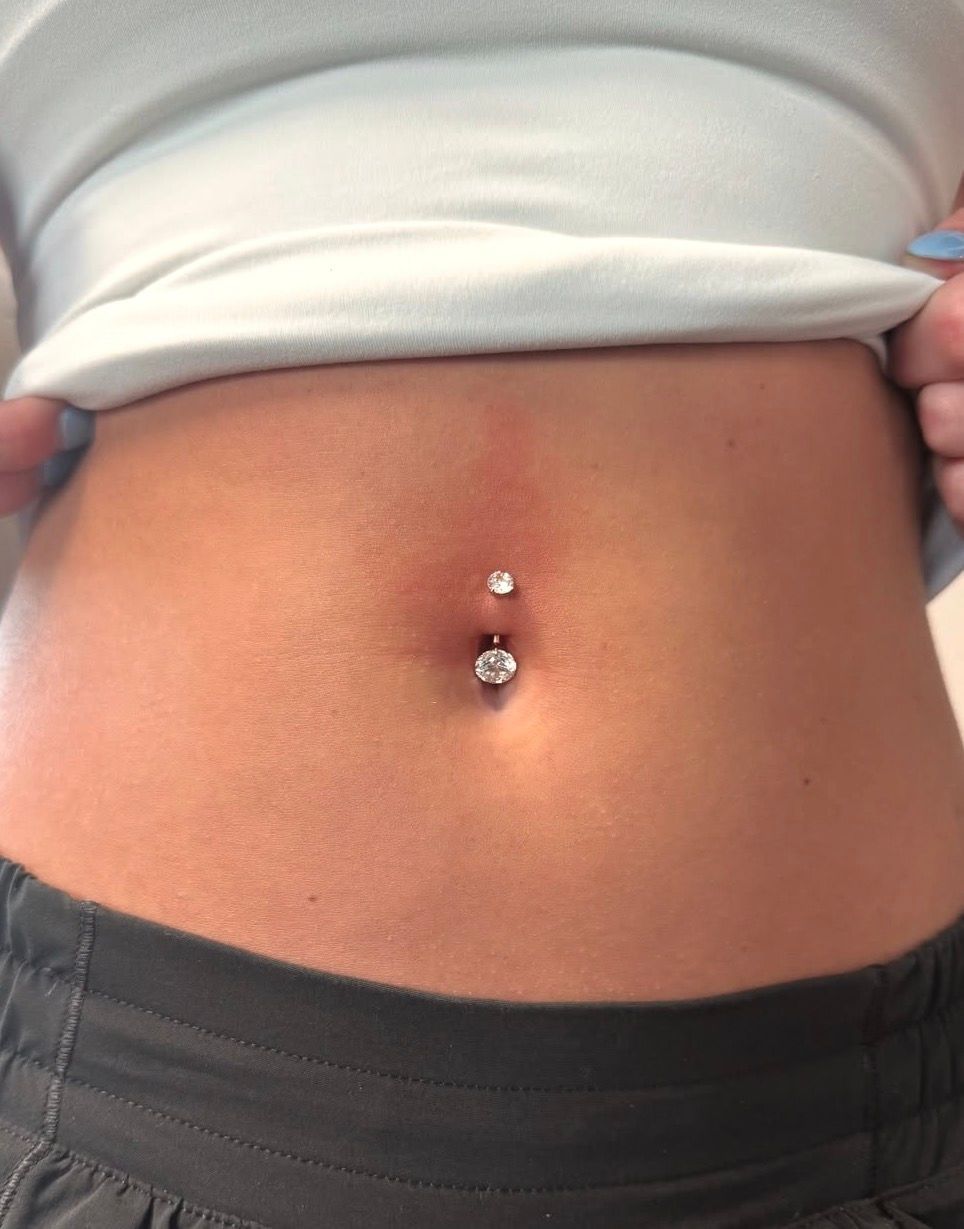 Close-up of a person's abdomen with a fresh navel piercing. Redness around the piercing, with two jeweled studs.