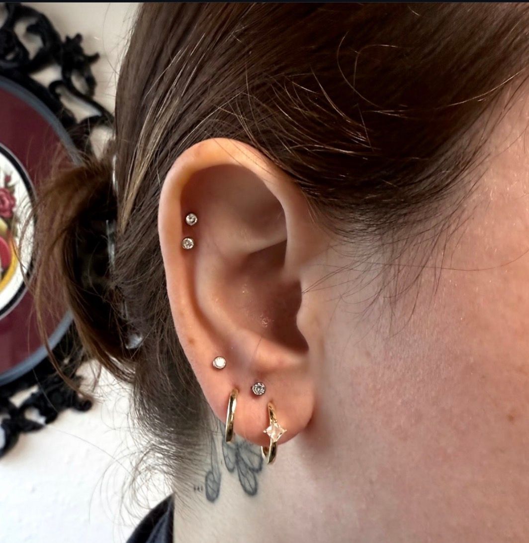 Close-up of ear with multiple piercings, including hoops and studs. Dark hair and a neck tattoo are also visible.