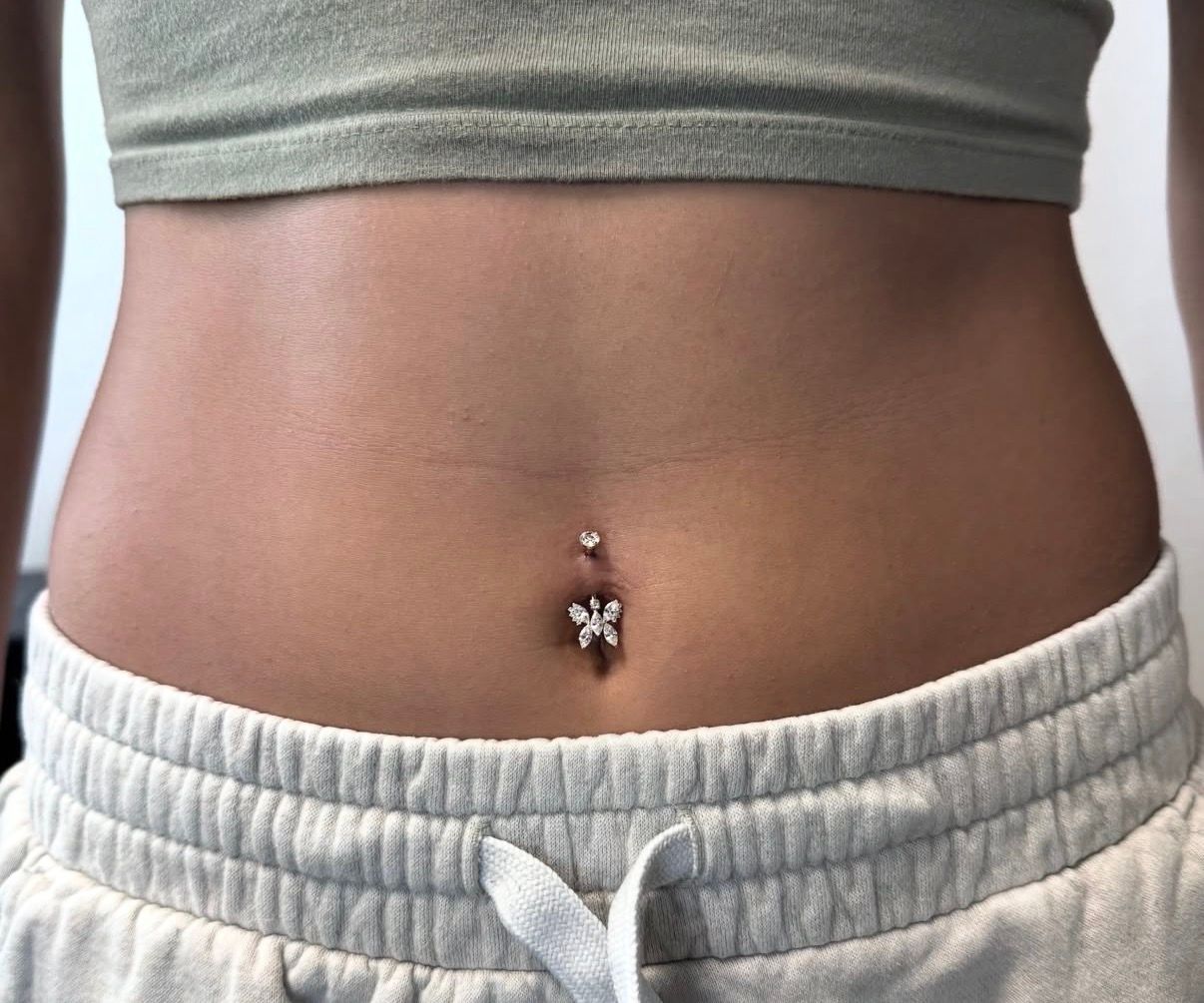 Close-up of a person's abdomen with a belly button piercing featuring a flower charm. They are wearing a crop top and sweatpants.