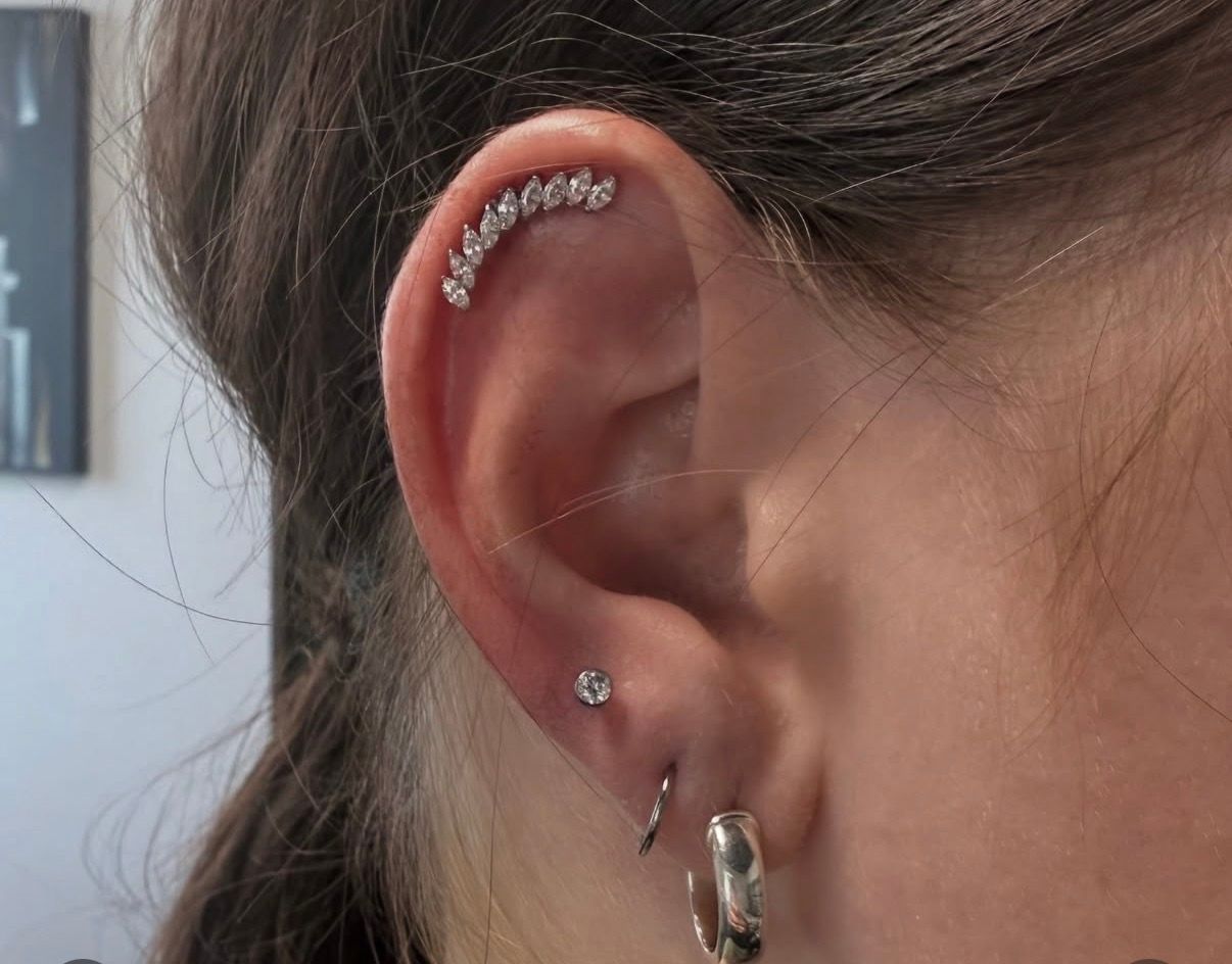 Ear with multiple piercings, including a curved cartilage piercing with small gemstones.