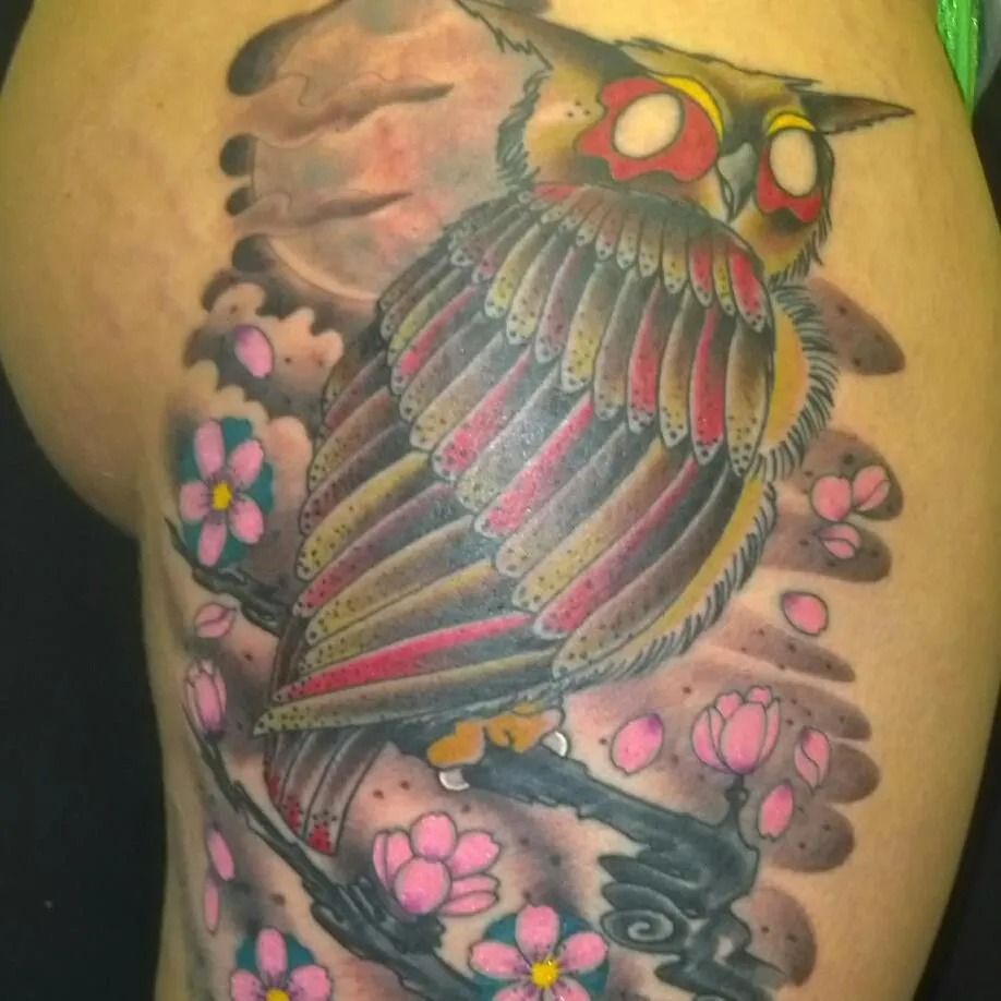 Tattoo of an owl perched on a branch, with cherry blossoms and cloudy sky background.