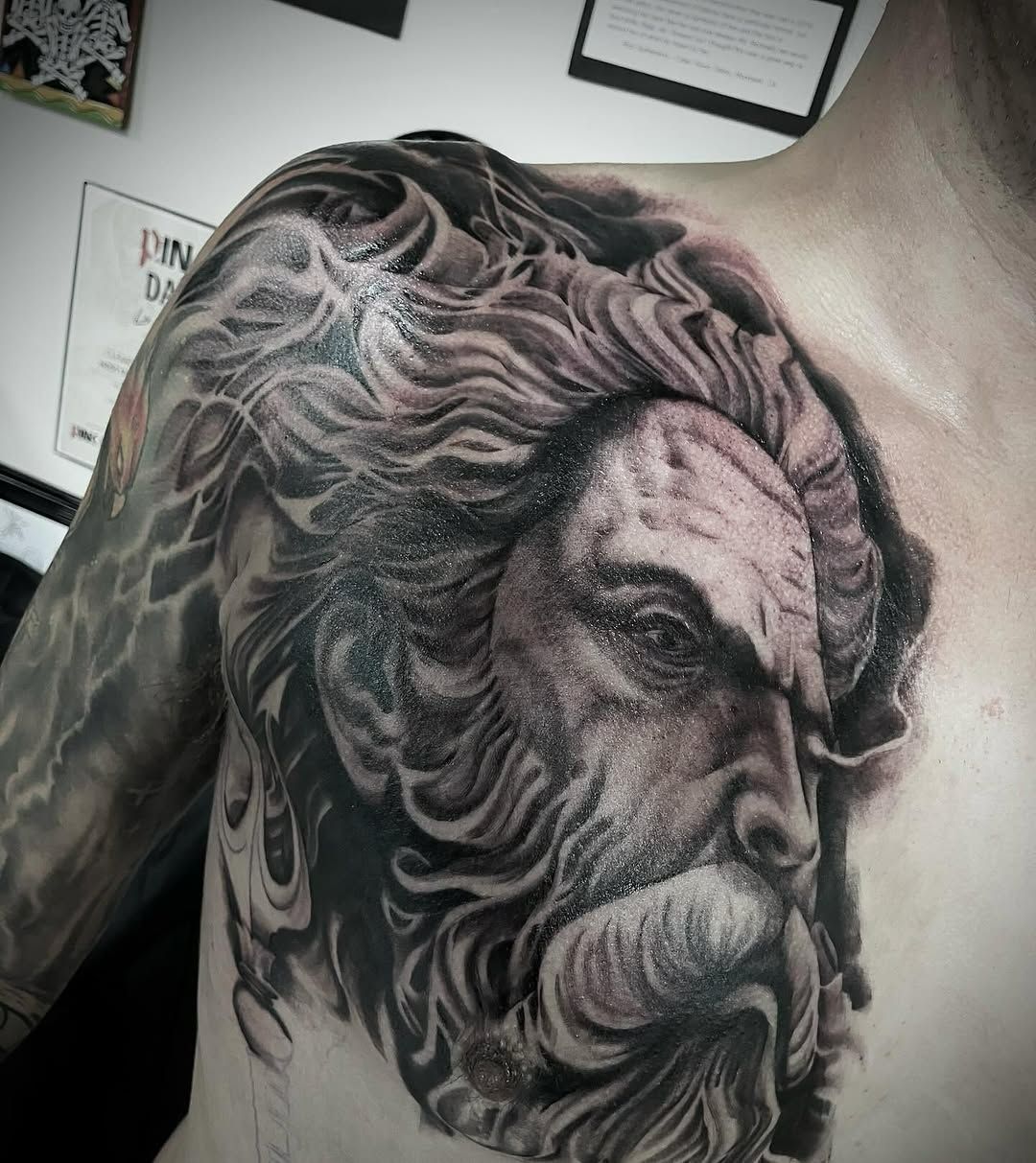 Tattoo of a stern-faced bearded man, likely a Greek god, on a person's chest and shoulder. Gray-scale.