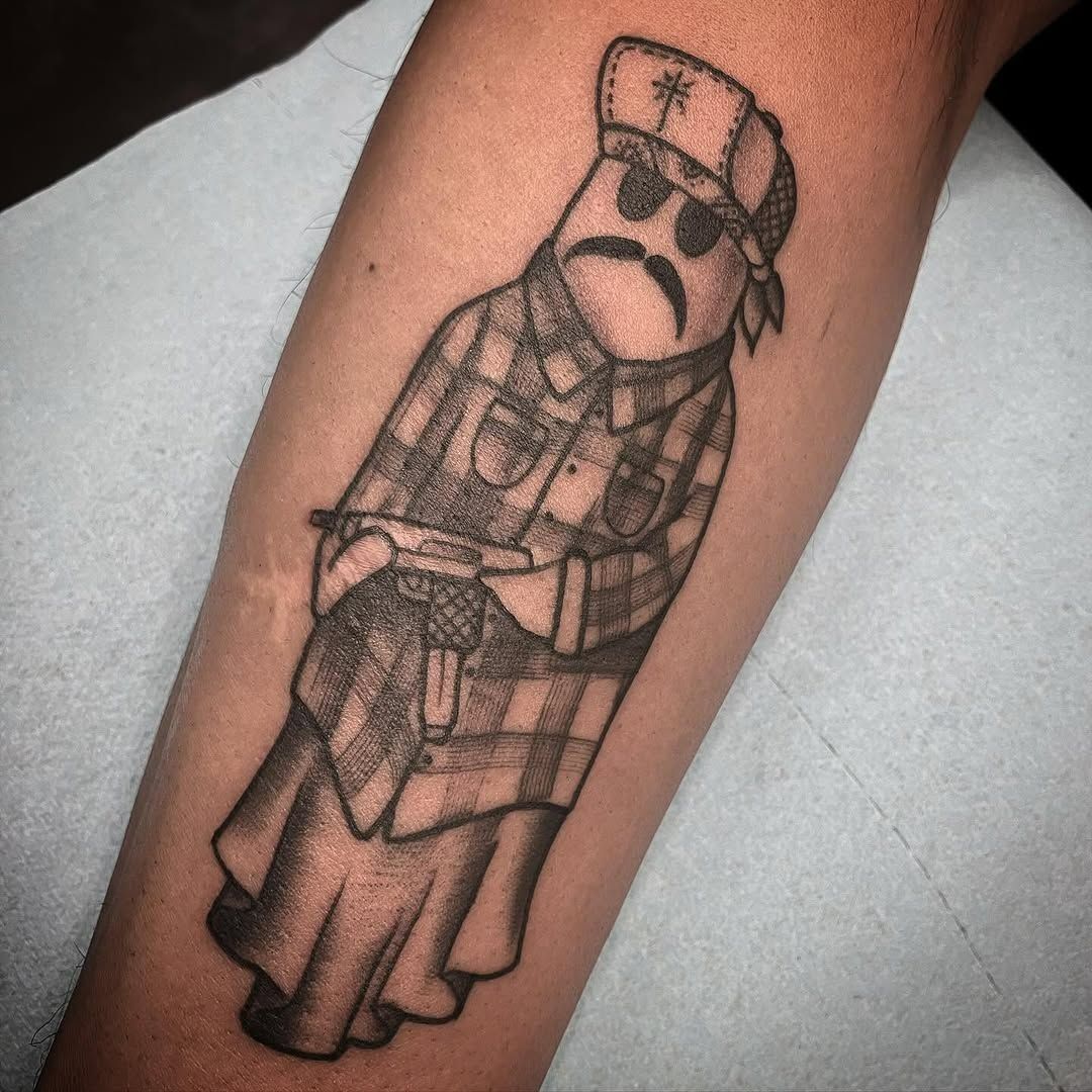 Tattoo of a sad-faced cartoon figure in plaid clothing and hat, holding a gun, on an arm.