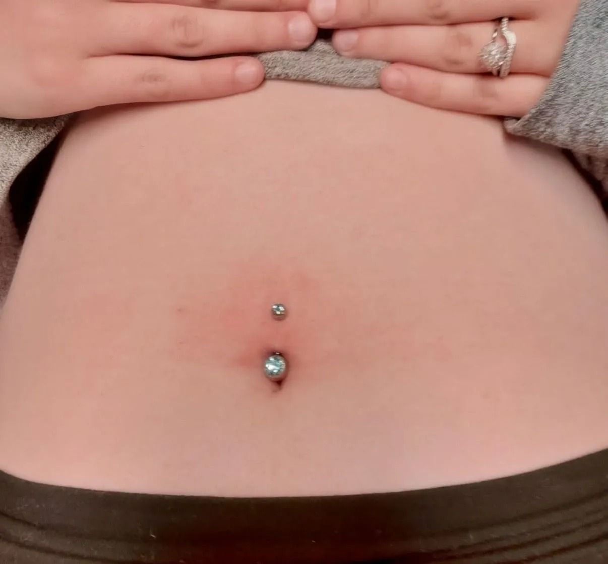 Close-up of a person's belly with a silver belly button piercing. The skin is pinkish, and the person’s hands are visible.