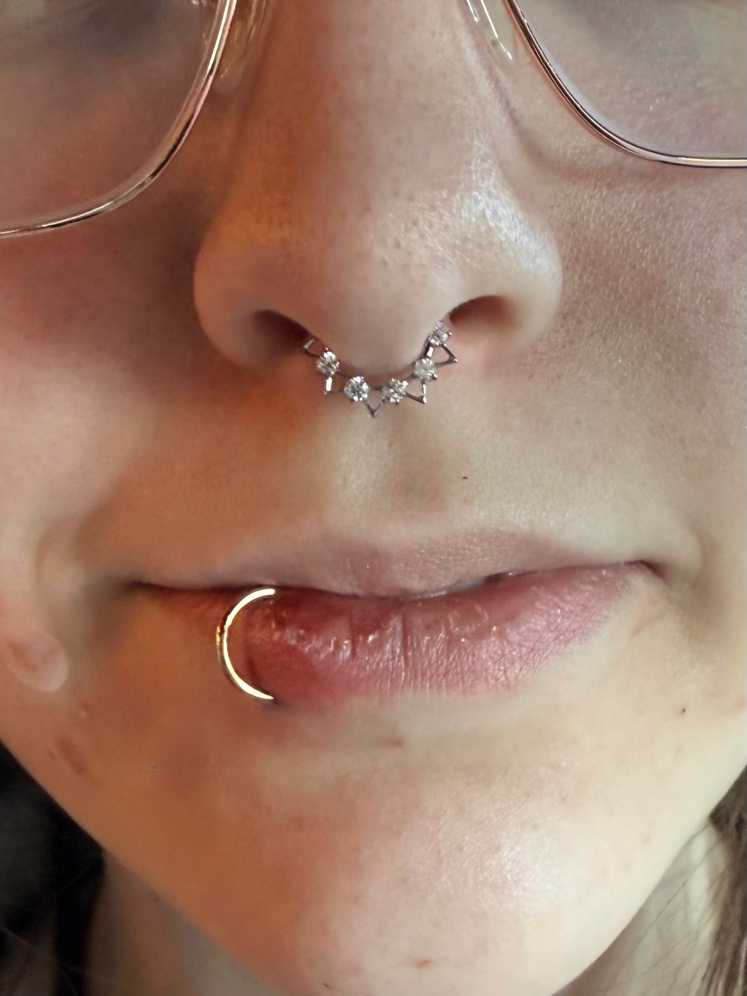 A close-up of a person's lower face showing a jeweled septum clicker and a gold lip ring.