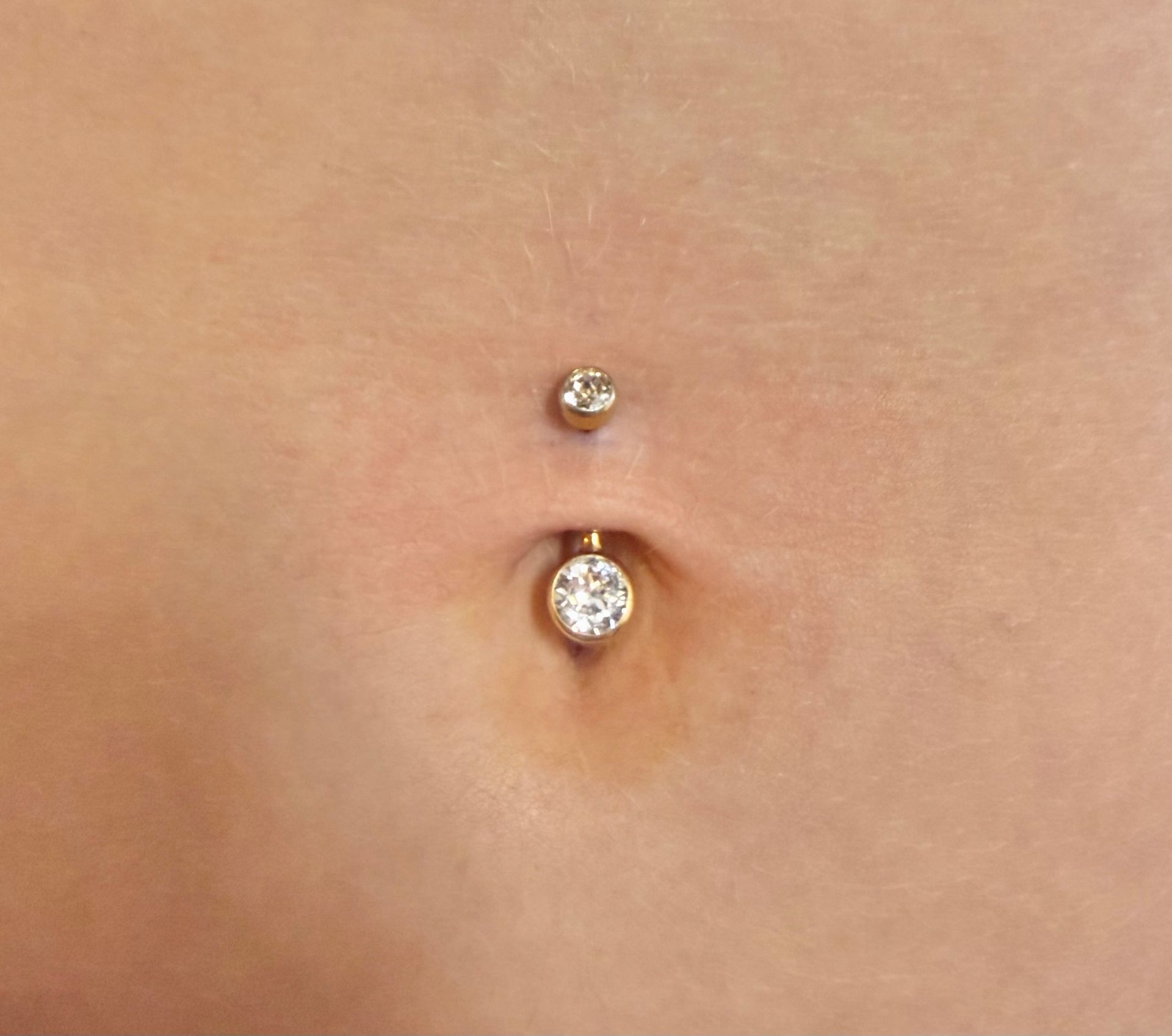 A navel piercing featuring a silver barbell with a small top ball and a larger, clear jeweled bottom charm.