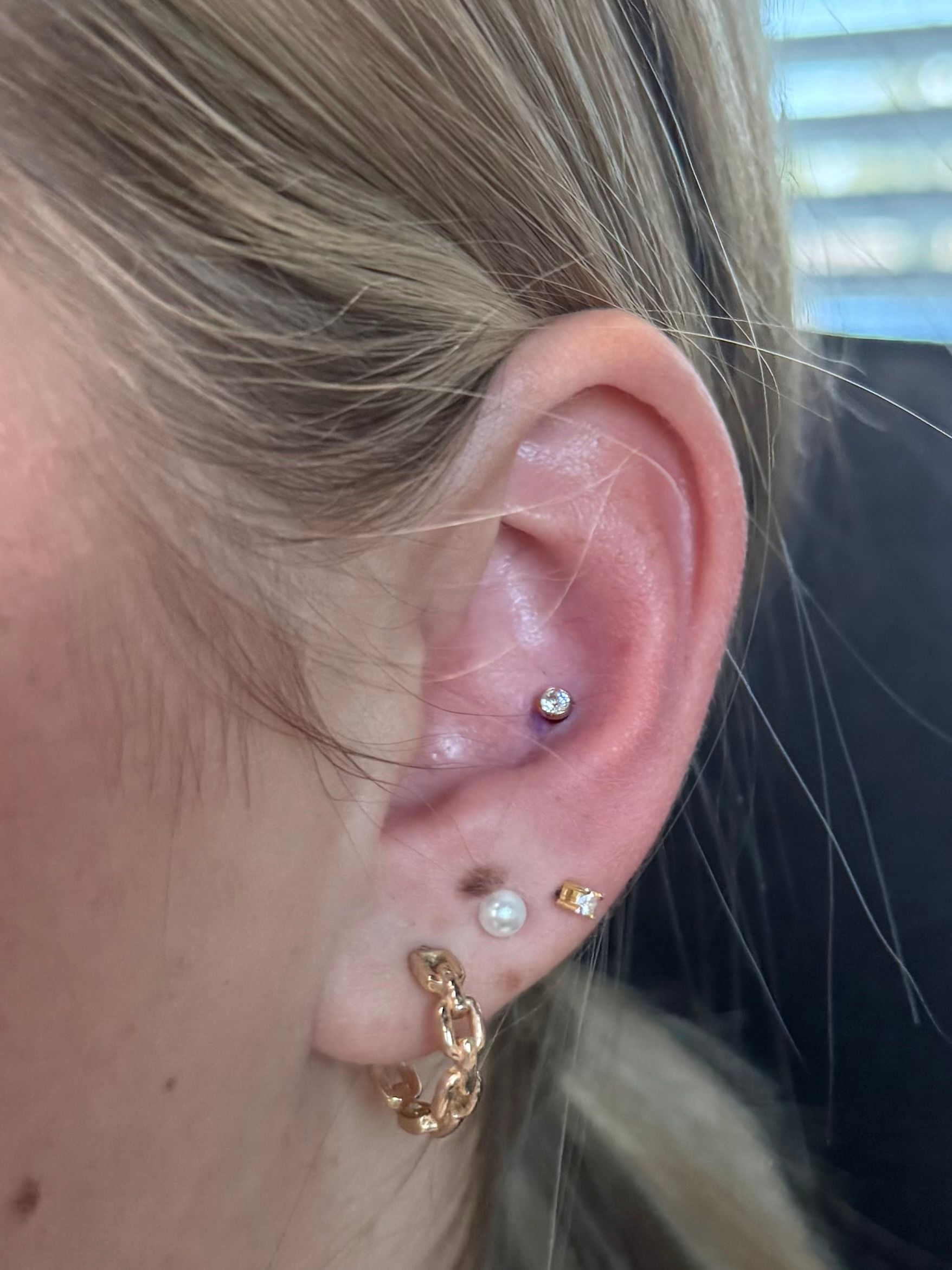 Close-up of a right ear with a conch piercing featuring a silver stud, plus two lobe piercings with a hoop and stud.
