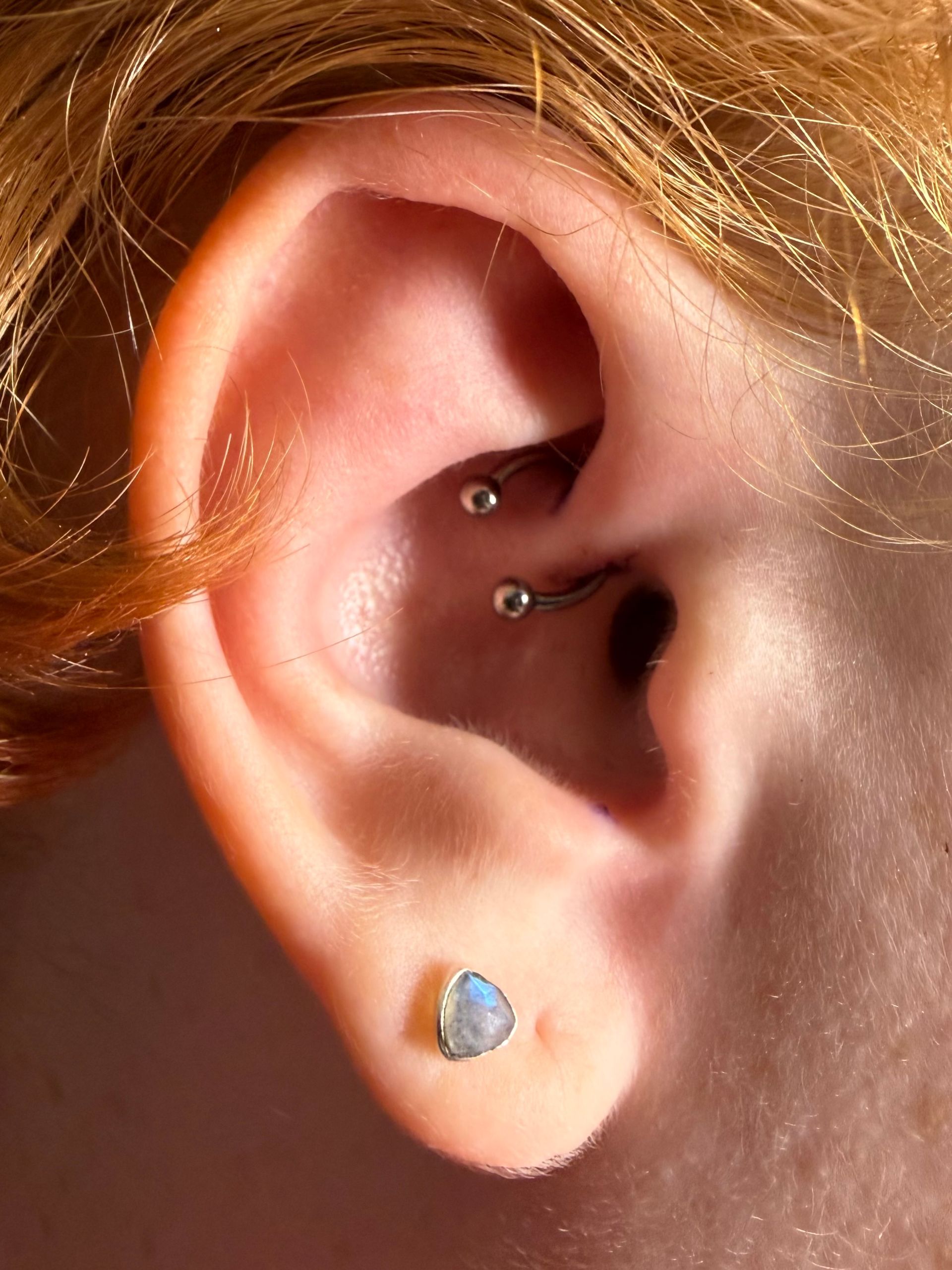 Close-up of a human ear with a circular barbell rook piercing and a triangular, blue-hued stone lobe earring.