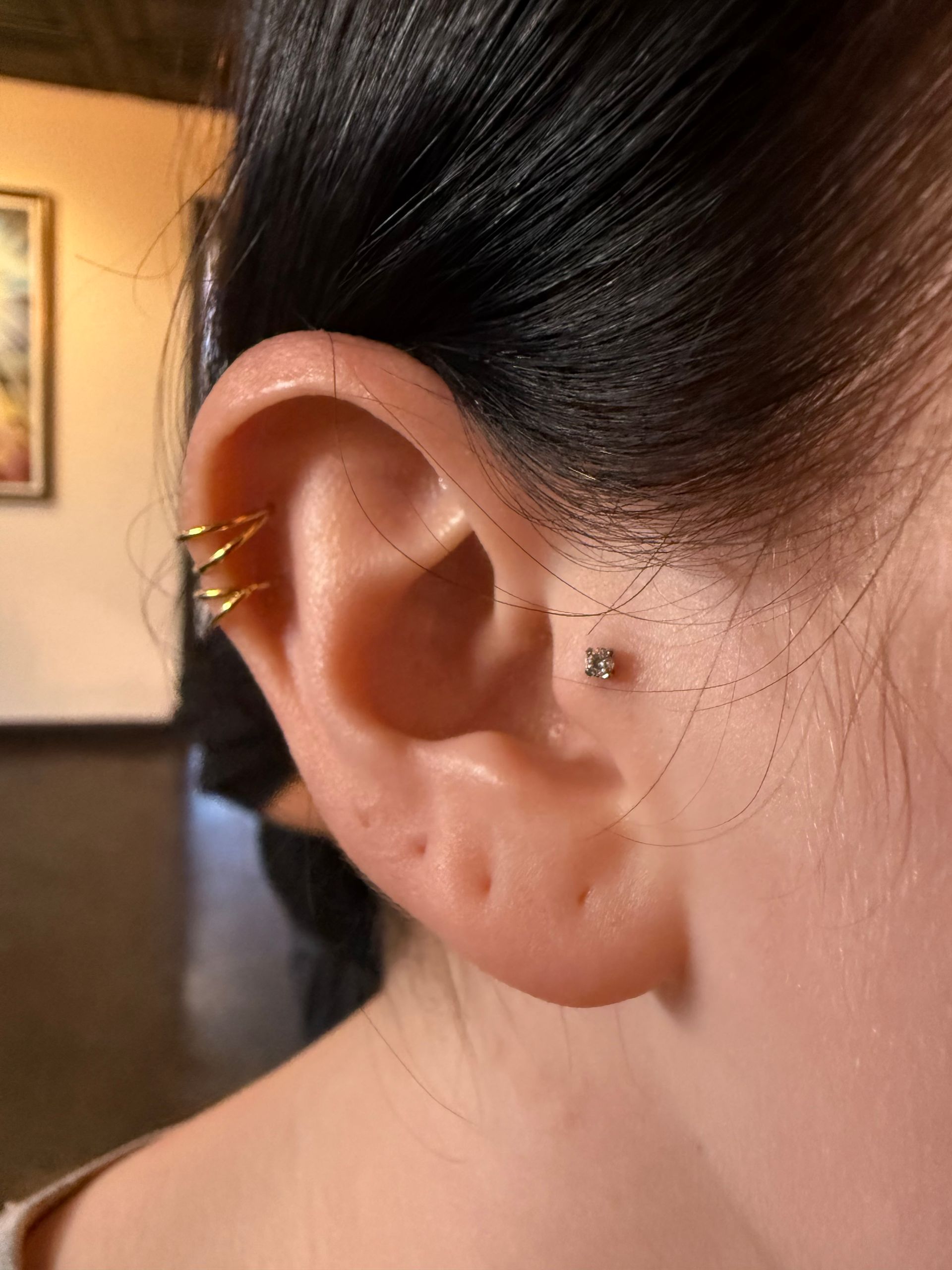 Close-up of a person's ear featuring a gold triple-hoop helix piercing and a silver stud tragus piercing.