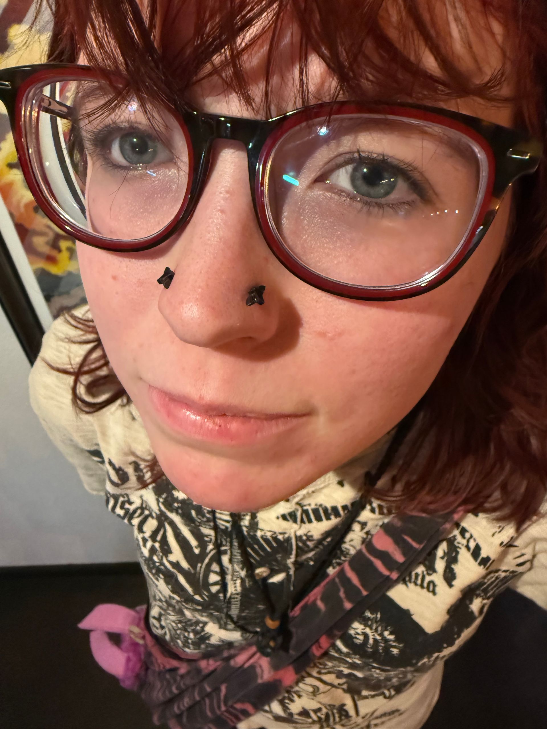 A person with red hair and glasses, featuring two black stud piercings on their nose, looking directly at the camera.