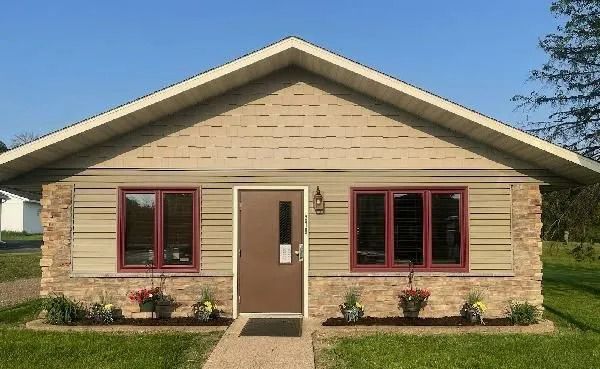 Small tan building with stone accents, brown door, two red-framed windows, flower boxes, and a grassy lawn.