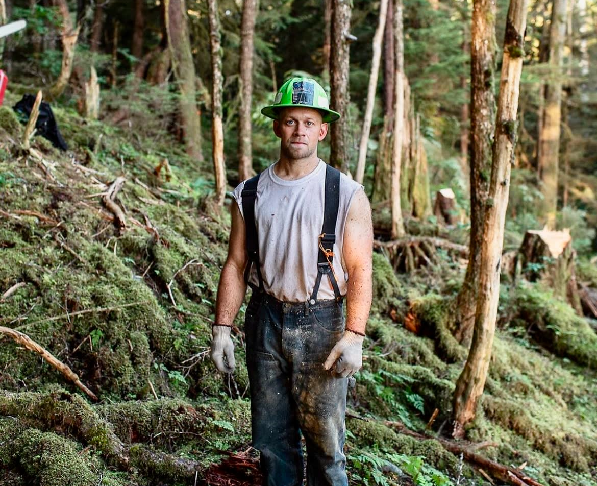A man wearing a hard hat and suspenders is standing in the middle of a forest.