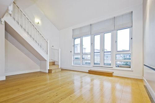 Bright empty living room with white walls, wooden floor, staircase, and large windows.