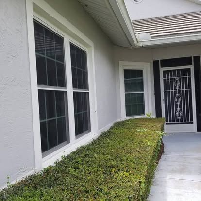 Front entrance walkway beside a white stucco house with trimmed hedges and a black security door