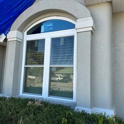 White arched window with shutters on a beige stucco house, framed by shrubs and blue sky.