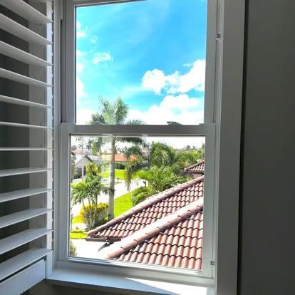 Open window with white shutters overlooking a sunny tropical neighborhood and red-tiled roofs