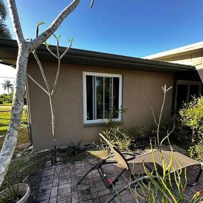 Small beige house exterior with a window, tree trunks, and a patio table and chairs in the foreground