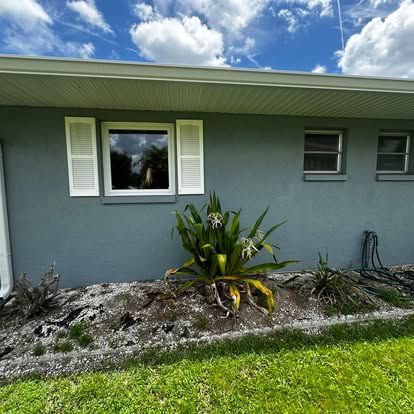 Gray house exterior with white shutters, green lawn, and a small tropical plant bed under a blue sky