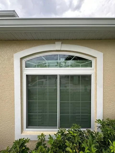 A beige house exterior with an arched white-trimmed window and green shrubs below.