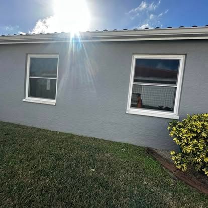 Gray house exterior with two windows, bright sun flare, green lawn, and a yellow shrub on the right