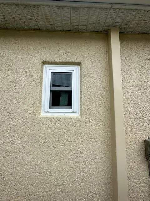 Small white-framed window set in a beige stucco exterior wall under an overhang.