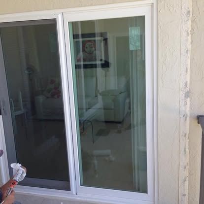 Sliding glass patio door with a dim reflection of a living room inside, and a hand holding cleaning spray in the corner
