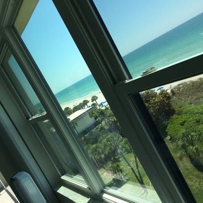 Tilted view through a window of a sunny beach, blue ocean, and green trees outside.