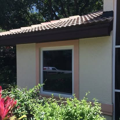 Small beige house exterior with a brown tiled roof and a window above green shrubs
