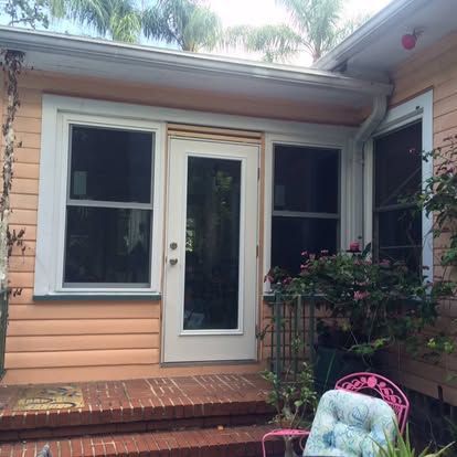 Small house entry with peach siding, white door, two windows, and a potted plant on a brick porch.