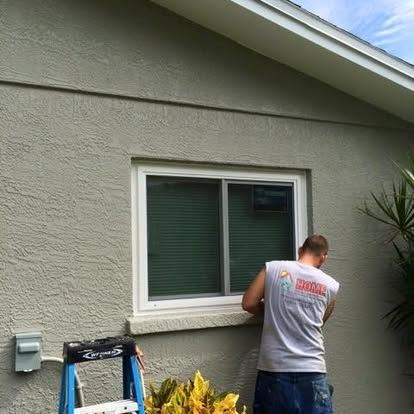 Person painting the exterior wall of a house beside a window and ladder.