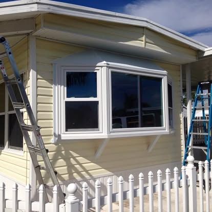 Mobile home exterior with white trim, bay window, and ladders beside a white fence