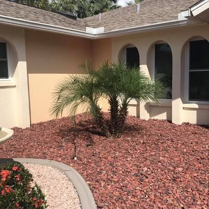 Landscaped front yard with a small palm tree, red rock mulch, and a stucco house with arched windows.
