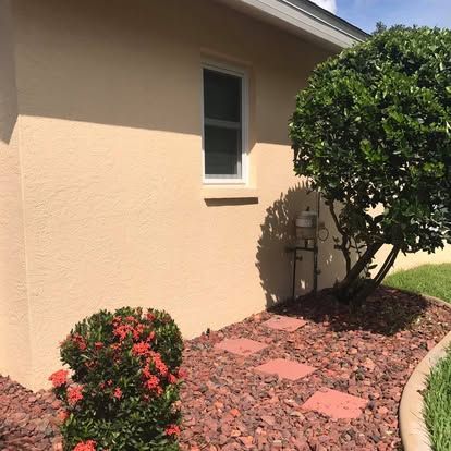 Landscaped side yard with a beige house wall, small windows, red stone mulch, and flowering shrubs.