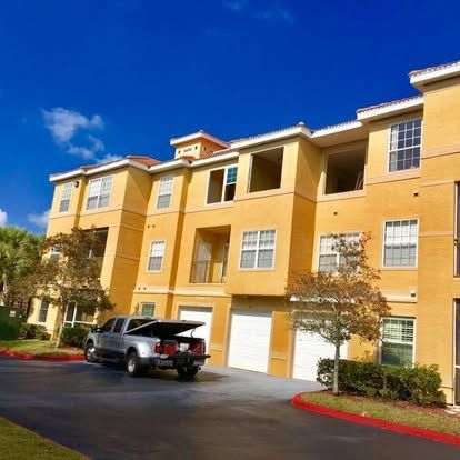 Three-story yellow apartment building with a parked SUV in front under a clear blue sky