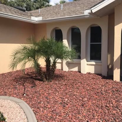 Front of a beige house with arched windows and a small palm tree in red rock landscaping