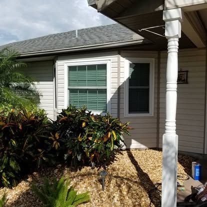 Front yard of a house with white siding, a window, shrubs, and a porch column in bright sunlight