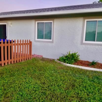 Single-story house exterior with green lawn, white stucco wall, and small orange fence by the windows