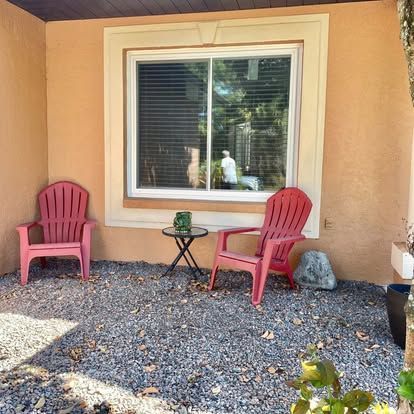 Two pink chairs and a small table beneath a window on a gravel patio, with a tan stucco wall background