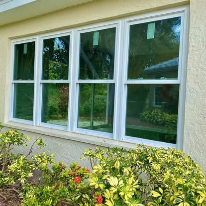 White-framed windows on a cream house wall above green shrubs and trees reflected in the glass