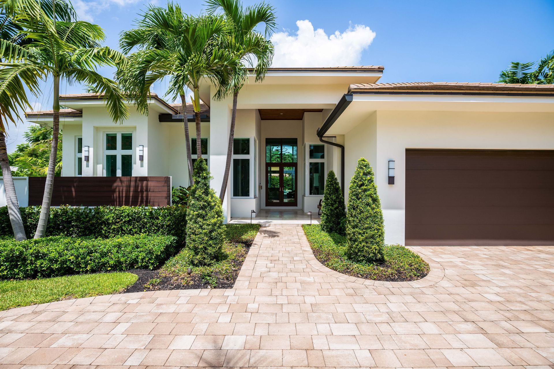 Modern single-story house with white exterior, palm trees, and a brick driveway leading to the front door