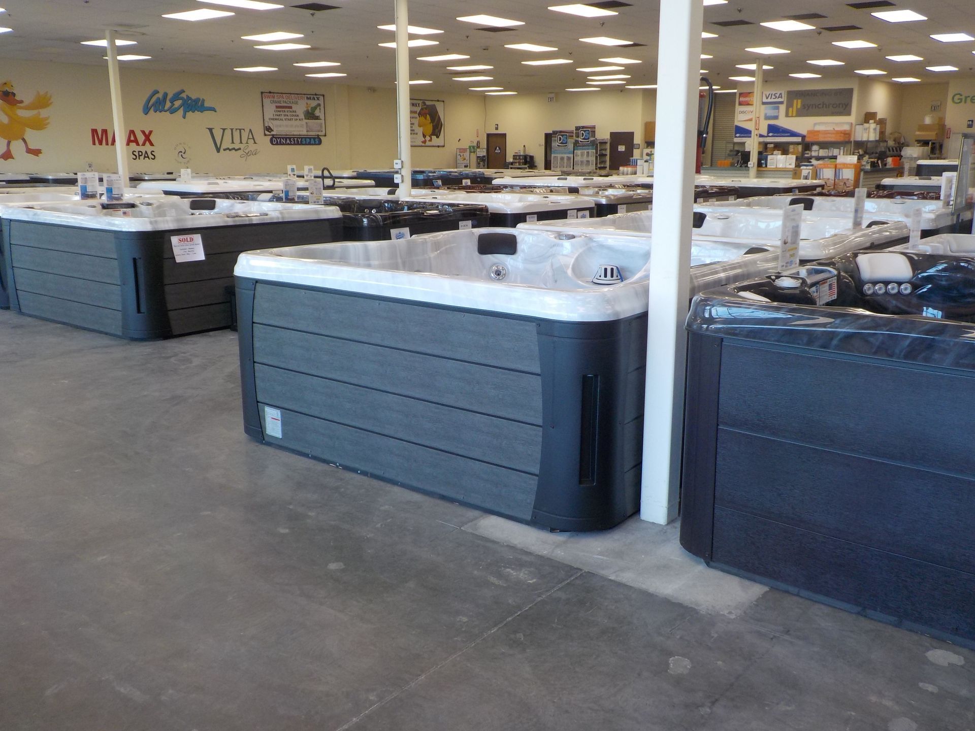 A showroom floor filled with rows of dark gray, rectangular hot tubs under bright indoor lighting.