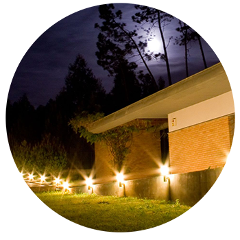 Night exterior of building with ground lights illuminating the brick and greenery, with trees and moon overhead.