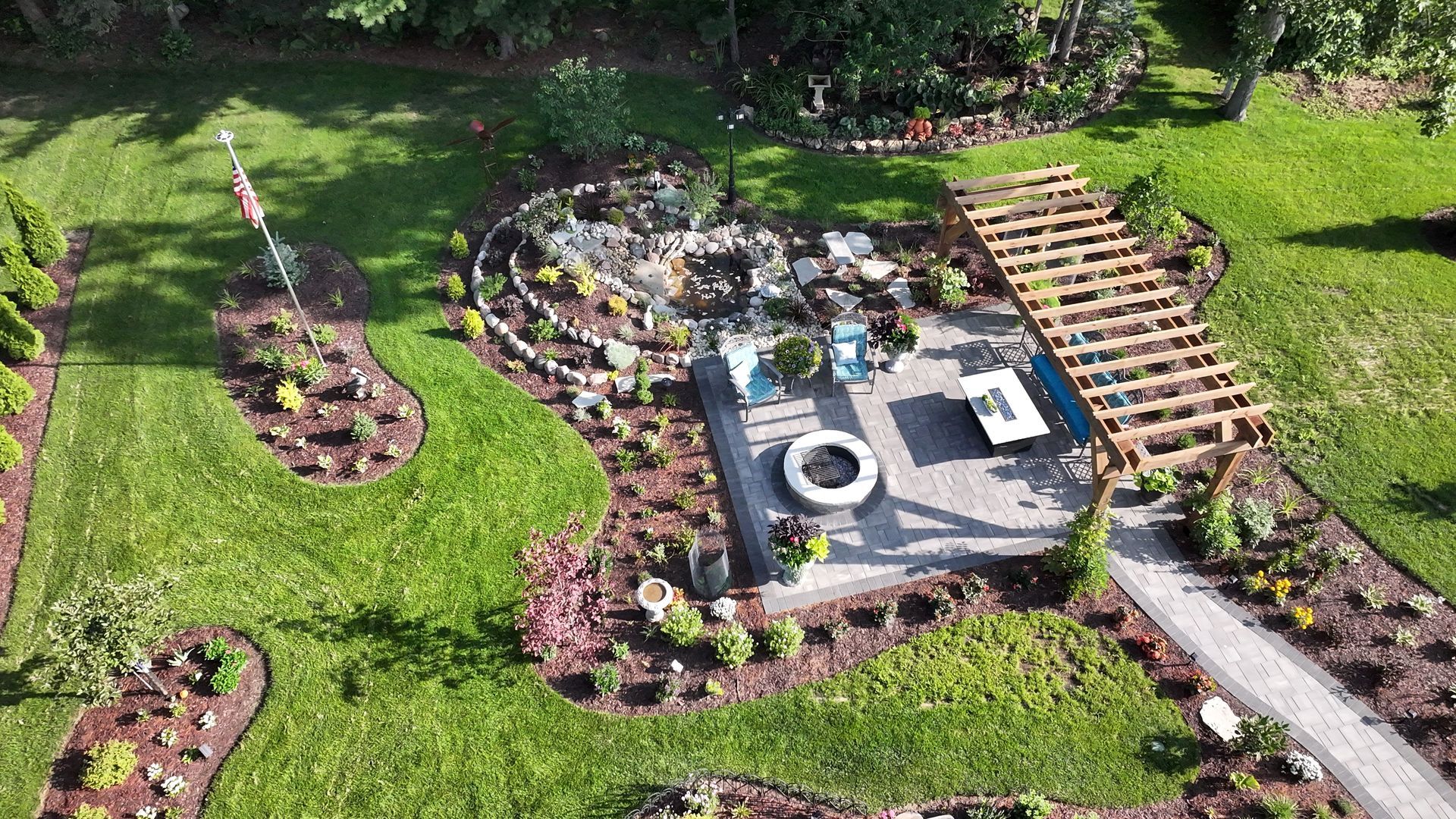 Aerial view of a landscaped backyard with patio, pergola, fire pit, and flag on a sunny day.