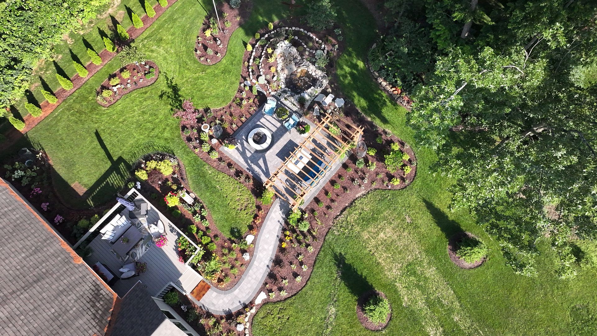Aerial view of a backyard with a stone patio, fire pit, pathway, and landscaped garden. Lush green lawn surrounds the area.