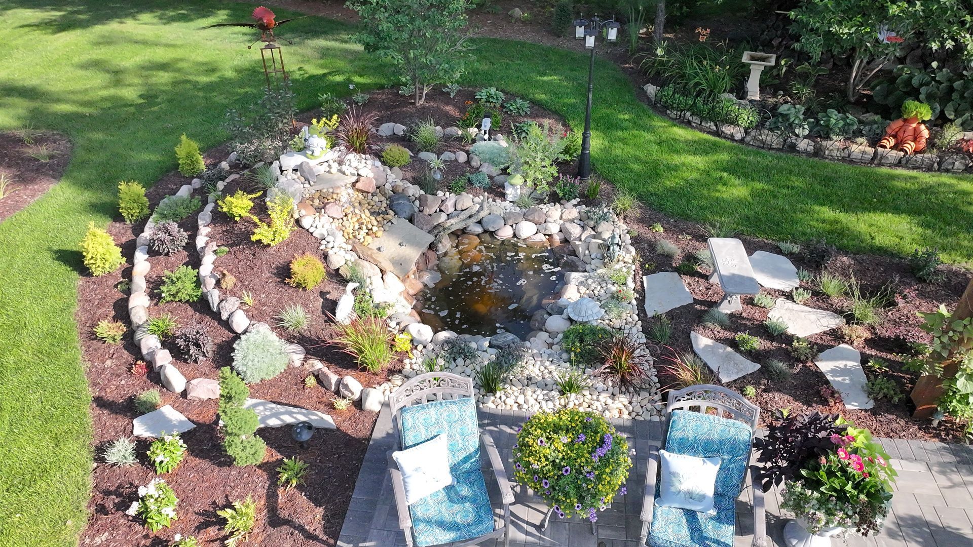 A well-landscaped garden with a water feature, stepping stones, and seating area, surrounded by green grass and plants.