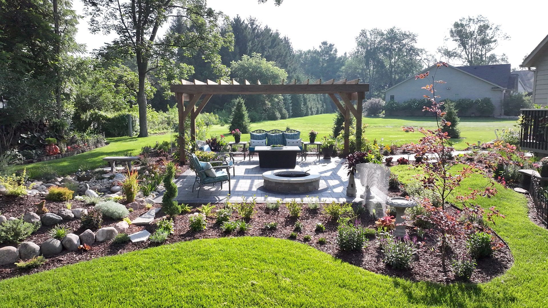 Backyard patio with pergola, fire pit, and seating surrounded by lush green lawn and landscaping.