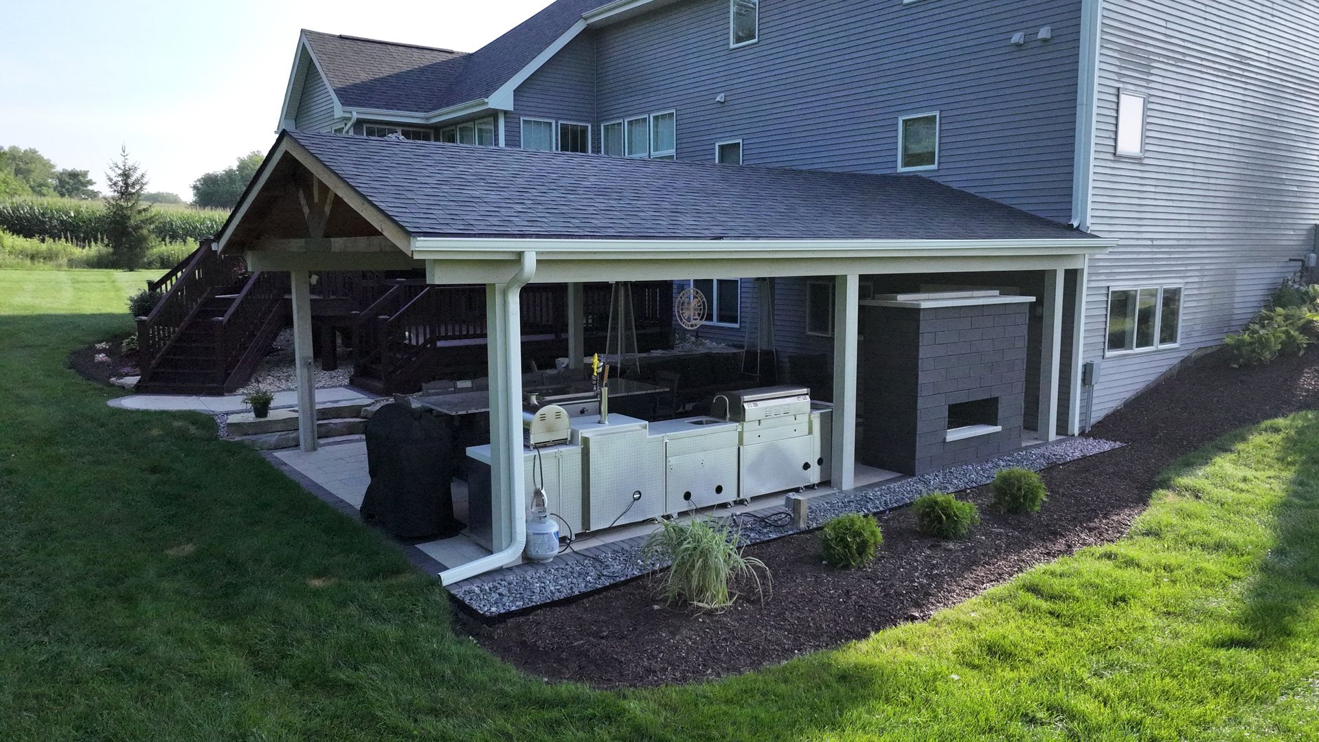 Backyard patio with outdoor kitchen, fireplace, and greenery. House in background.